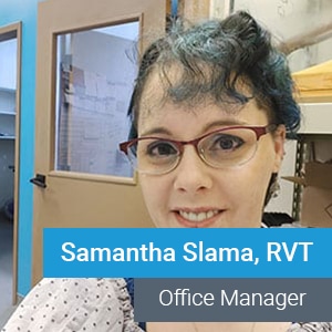 A woman with glasses and short, curly, blue-tinted hair smiles at the camera in an office. Text on the image reads: “Samantha Slama, RVT - Office Manager.”.