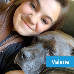 A woman with long brown hair smiles while cuddling a large gray dog on a couch. There is a blue label in the bottom right corner with the name "Valerie" on it.